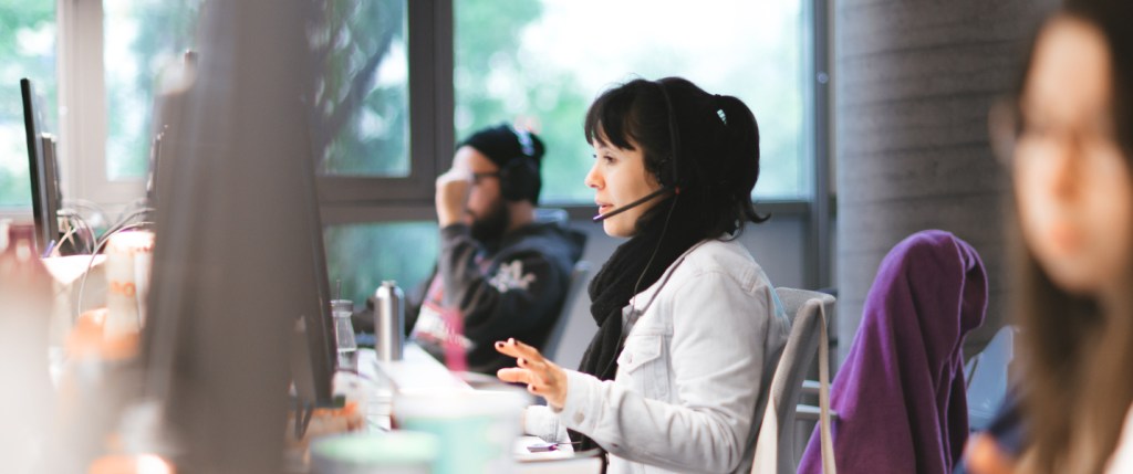 Três pessoas trabalhando no escritório do Nubank. O foco da foto está em uma mulher, jovem, atendendo clientes com um headphone