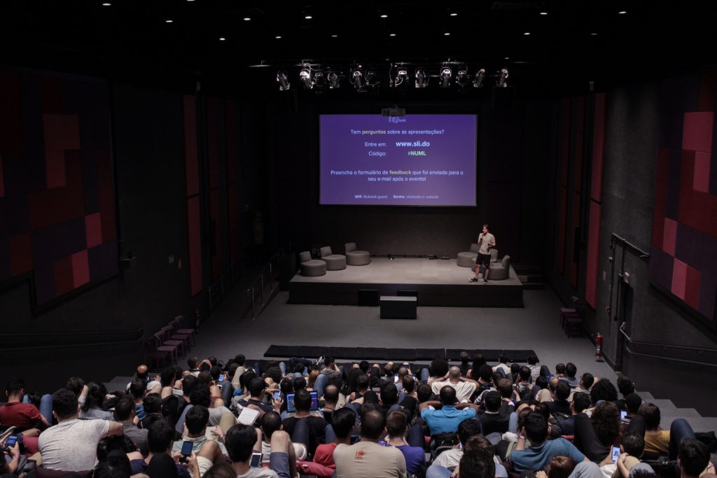 Encontro de Machine Learning no auditório do Nubank. A foto mostra os espectadores de costas e, ao fundo, um palestrante no palco