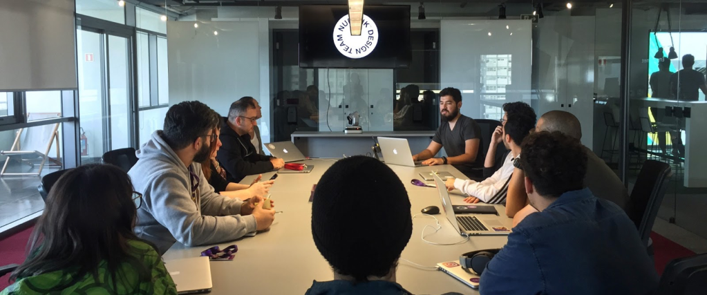 Sala de reunião com grupo de pessoas reunidas em torno de uma mesa comprida. Uma TV, pendurada no teto, mostra o símbolo do Design no Nubank