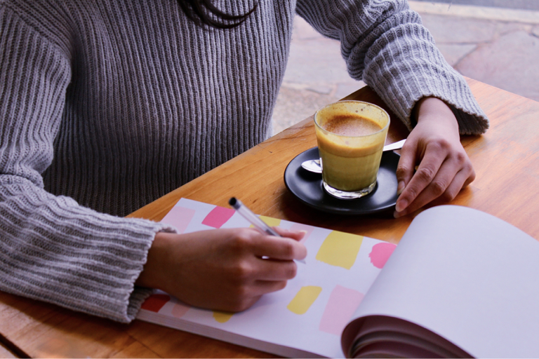 Pessoa sentada em uma mesa, comum café e um bloco de anotações