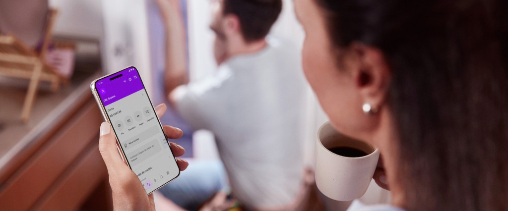 Imagem de uma mulher branca de cabelos escuros segurando o celular na tela do app Nubank em uma mão e uma xícara de café na outra. Ao fundo, um homem branco abaixado consertando algo na parede.