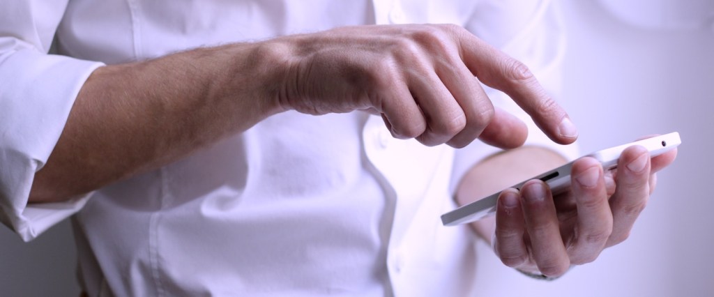 Estou negativado. Posso abrir uma conta digital? Um homem segurando o celular com a mão esquerda e com o indicador direito quase tocando a tela.