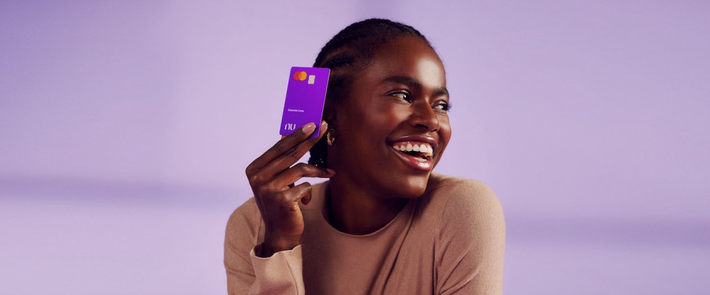 Foto de uma mulher negra segurando um cartão Nubank. O fundo é lilás.