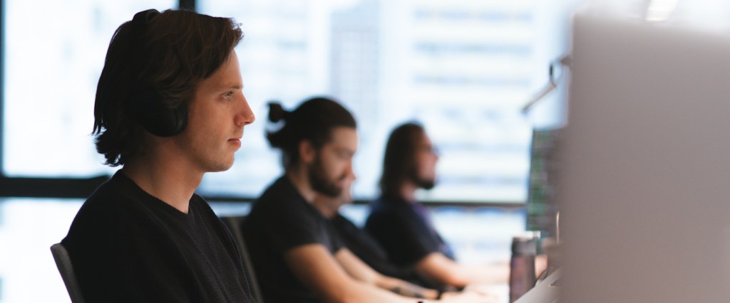 Machine learning: foto de um homem trabalhando. Ele veste preto, usa fones de ouvido e está digitando no teclado. Mais ao fundo, outros dois homens também estão trabalhando em seus computadores.