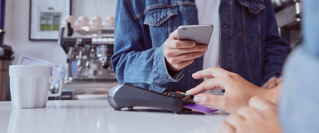 Nubank: foto dentro de uma cafeteria. Sobre o balcão branco, uma pessoa está digitando a senha na maquininha de cartão. O cartão de crédito Nubank está inserido na máquina. Atrás do balcão, um homem de camiseta branca e jaqueta jeans está com o celular na mão esquerda.