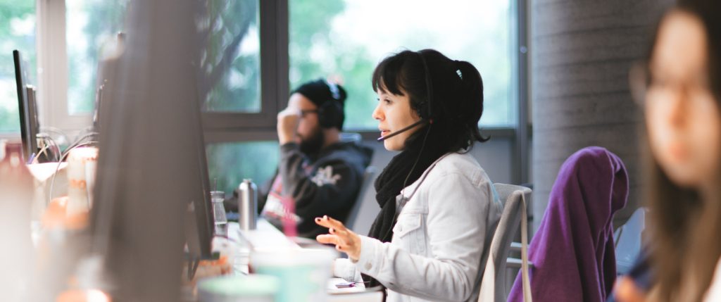 Atendimento Nubank: Três pessoas trabalhando no escritório do Nubank. O foco da foto está em uma mulher, jovem, atendendo clientes com um headphone