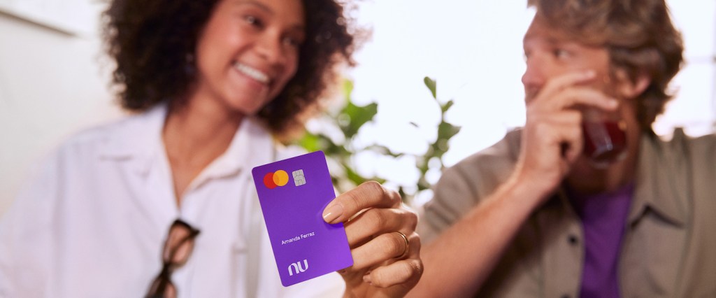 Imagem de uma mulher negra sorrindo, segurando o cartão Nubank. Ela está usando uma camisa branca e tem óculos de sol pendurado nela. Ao lado, um homem loiro, com uma camiseta roxa e uma camisa em tom de verde por cima. Os dois parecem se olhar.