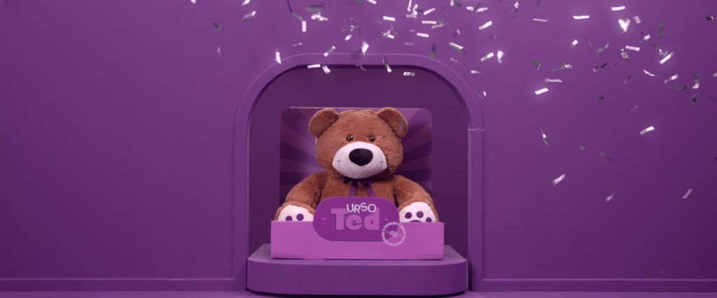 TED Bear: ursinho do Nubank em uma caixa e cenário roxo com chuva de prata caindo sobre ele