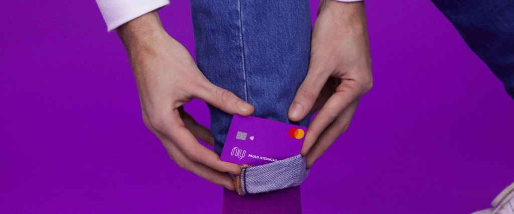 Produtos Nubank: uma pessoa vestindo uma calça jeans e tênis branco, sobre um fundo roxo, segura seu cartão Nubank do lado de sua perna.