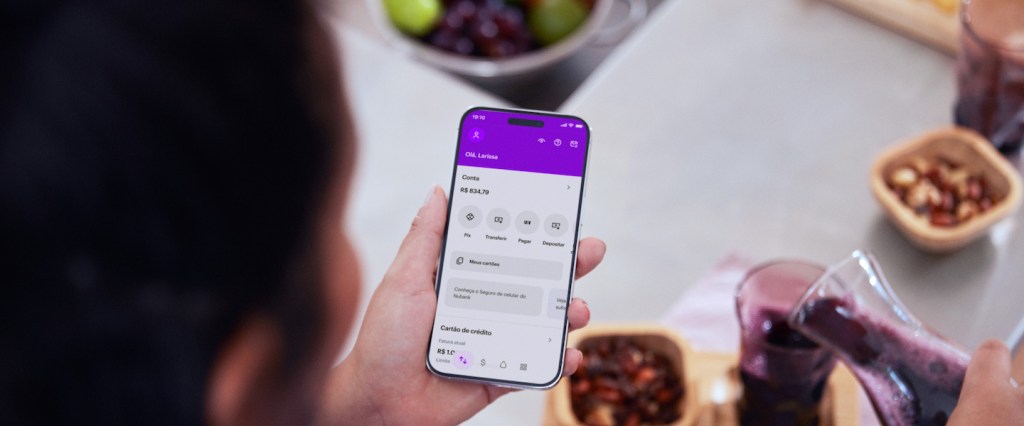 Imagem de uma mulher de costas, de cabelo cacheados, com um celular na mão e na tela o app do Nubank. Ela está colocando suco de uva no copo de vidro.