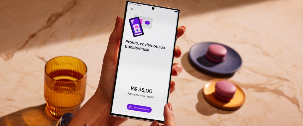 Como fazer TED no Nubank? A imagem mostra a mão de uma pessoa negra segurando o celular com a tela do app do Nubank após realizar uma transferência no valor de trinta e oito reais. A primeira mensagem na tela do app é: 