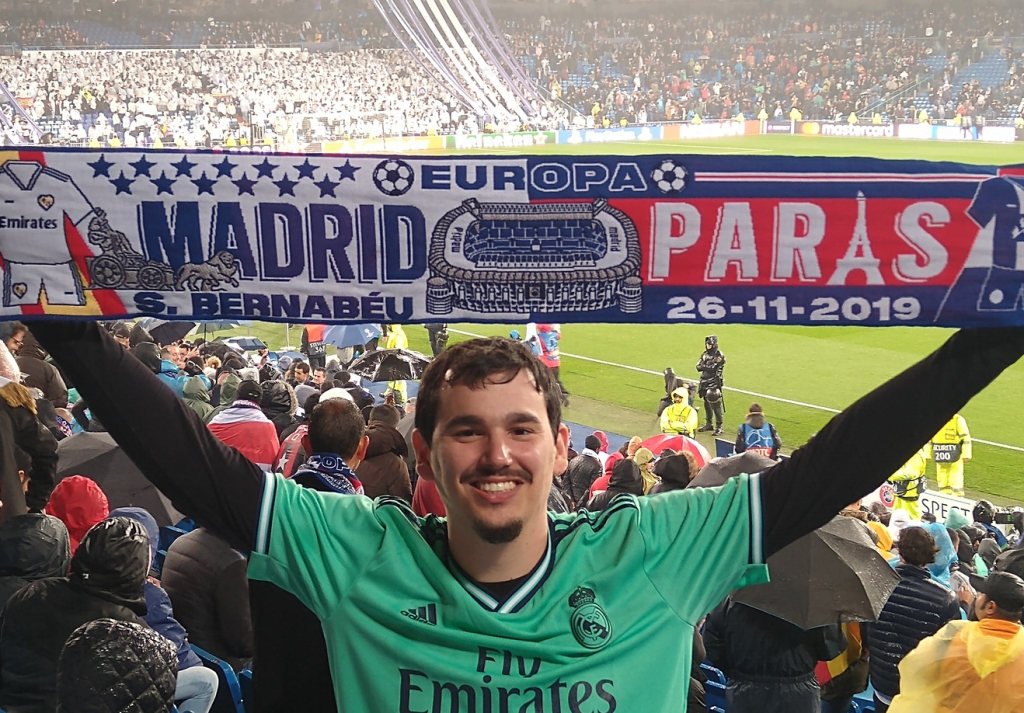 Arthur no jogo do Real Madrid, vestindo a camisa do time e segurando uma faixa de torcedor do time espanhol.