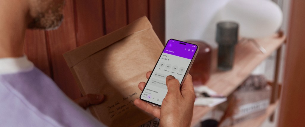 Imagem de um homem branco de meia idade segurando um pacote de papel pardo em uma mão e na um celular, na tela do aplicativo do Nubank.