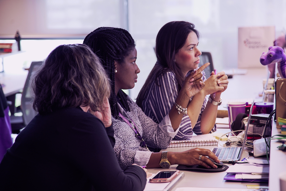 Três mulheres sentadas lado a lado  no escritório olhando para uma tela de computador. Uma delas aponta o dedo para frente.