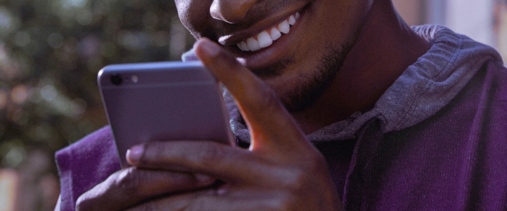 Jovem vestindo moletom roxo segura um celular em frente ao seu rosto enquanto sorri.