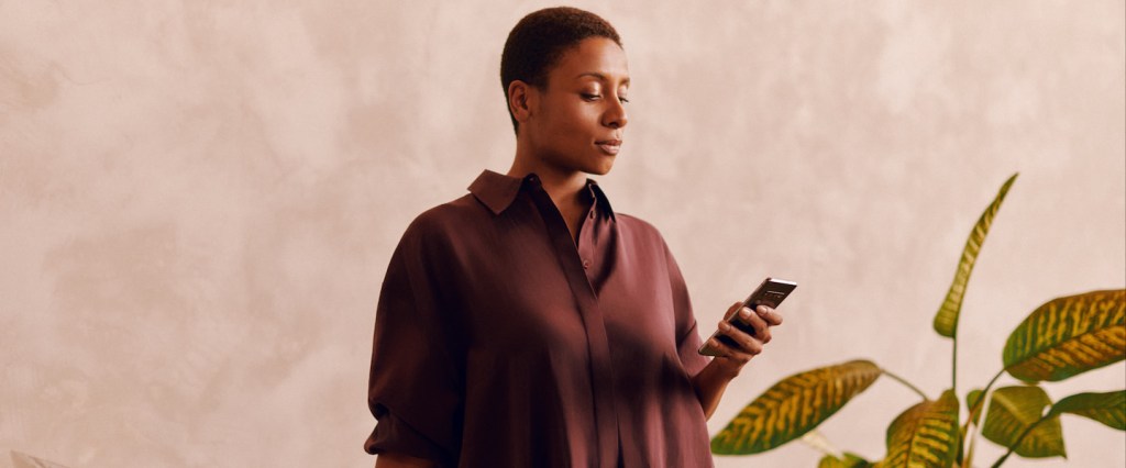 Como gerar boleto Nubank? Imagem mostra uma mulher negra vestindo um vestido marrom e olhando para a tela do celular que está segurando com a mão esquerda. Fundo com parede bege e uma planta comigo-ninguém-pode ao fundo do lado direito.