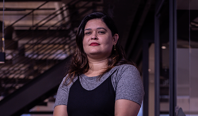 Mulheres Nubank: fotografia de Kete, líder técnica no Nubank, posando de pé no escritório. Ela veste macacão preto de alçar e camiseta cinza.