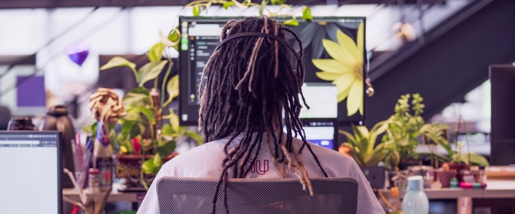 Atendimento Nubank: Jovem com dreads no cabelo sentado, de costas, em uma mesa de trabalho.