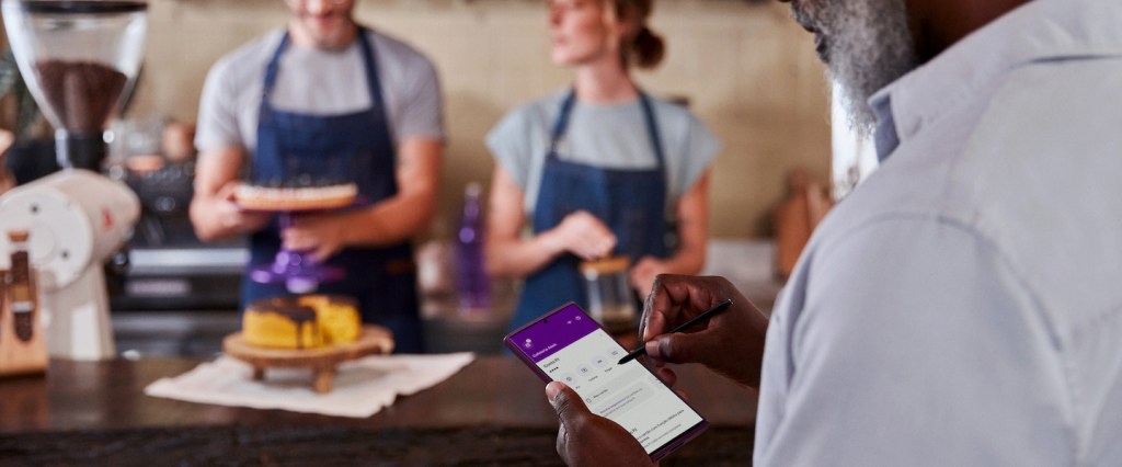 Conta PJ do Nubank: tudo o que você precisa saber sobre ela. Imagem mostra um homem negro em primeiro plano, ao lado direito da foto, olhando para o celular a tela inicial da conta PJ do Nubank. Ao fundo, um homem e uma mulher branca, ambos vestindo camiseta cinza e avental jeans, finalizando sobremesas com laços roxos.