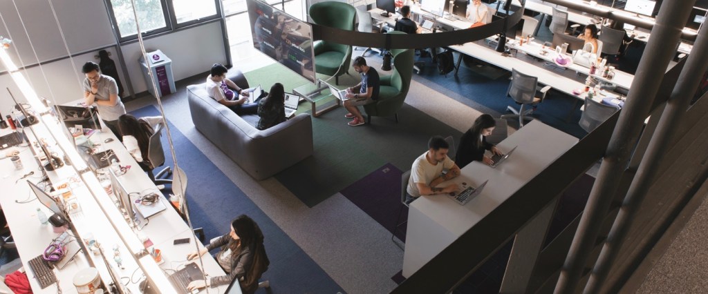 Picture taken from above shows an office, with green and gray carpet, open desks and people woking on computers