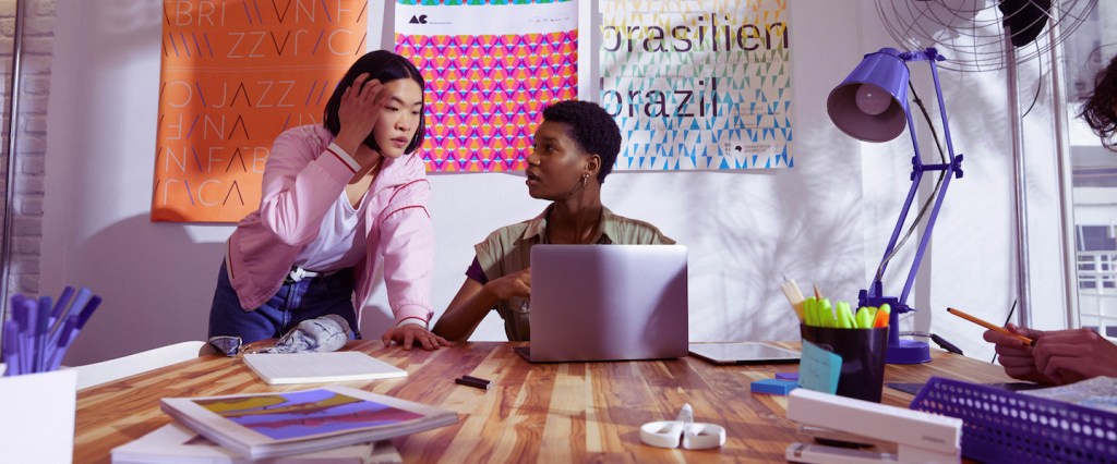 Boleto de cobrança e boleto de depósito: foto mostra duas jovens conversando em frente a uma parede de post its. Uma delas está sentada em uma mesa, com um computador aberto