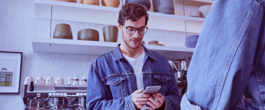 Atualização cadastral Nubank: imagem mostra jovem de óculos e jaqueta jeans, atrás de um balcão de café, olhando para o telefone