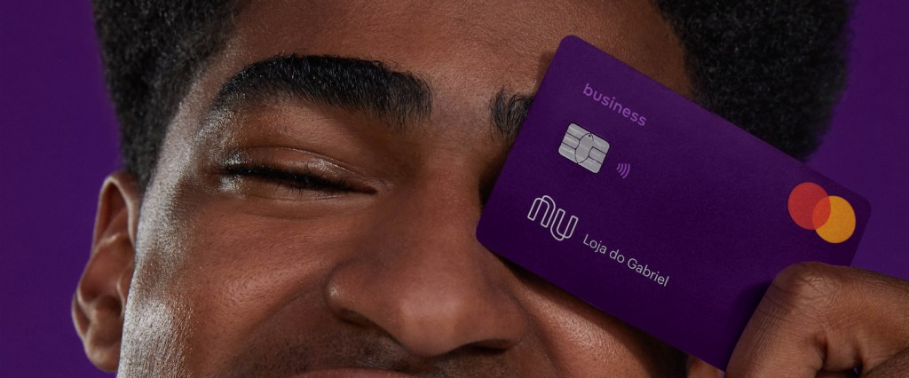 Como funciona o débito na conta PJ Nubank: foto em estúdio com fundo roxo de um homem sorrindo segurando o cartão PJ em frente ao olho esquerdo