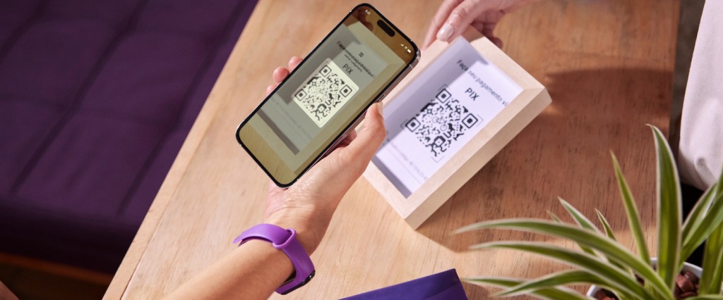 Pix na conta PJ do Nubank: tudo o que você precisa saber. Imagem mostra a mão de uma pessoa branca, usando um relógio roxo, seguranco o celular apontando a câmera para o código QR code impresso em uma moldura que outra pessoa está segurando. Ao fundo, um balcão de atendimento de madeira e uma planta.