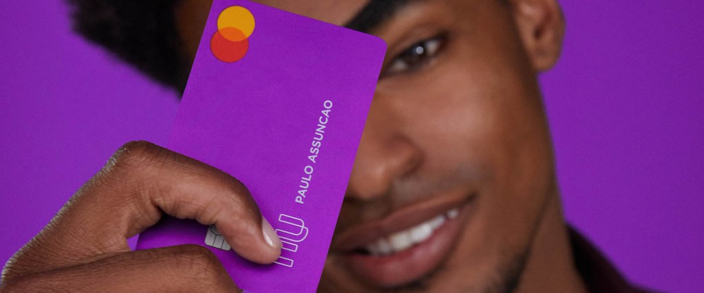 4 dúvidas da internet sobre o Cartão Nubank: fotografia de estúdio, no fundo roxo, de um homem segurando o cartão Nubank em frente ao rosto.