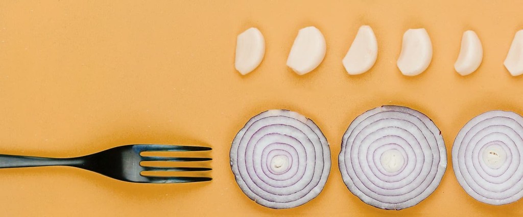 rodelas de cebola roxa e dentes de alho perto de um garfo, em um fundo amarelo