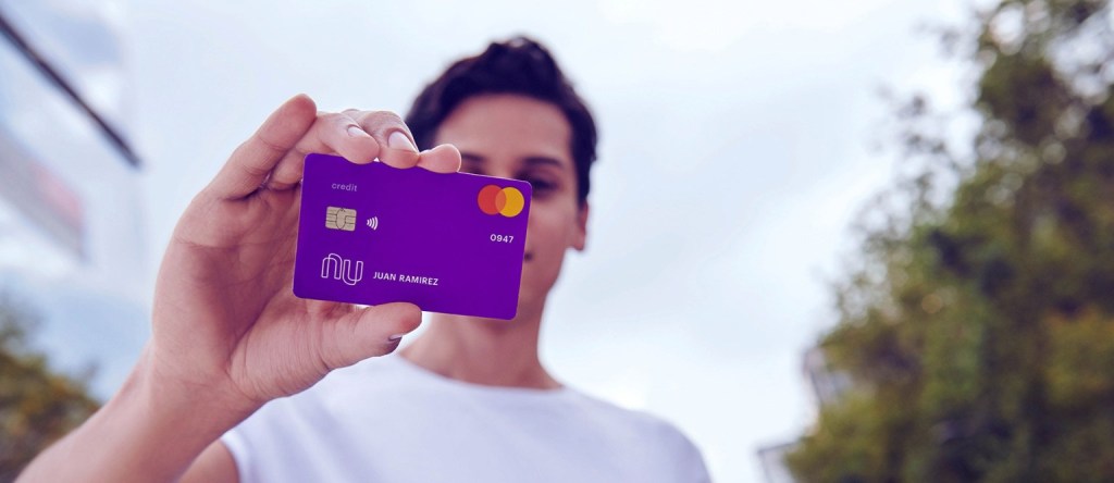 Jovem de camiseta branca segurando um cartão de crédito Nubank, roxo, na frente do rosto