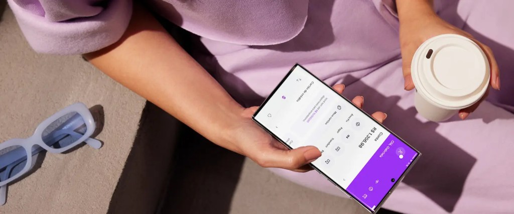 Nubank login: passo a passo para entrar no app e no site. Imagem de uma pessoa segurando um celular com a tela inicial do app do Nubank na mão direita. Na mão esquerda, um copo de café para viagem. A pessoa tem pele branca e veste uma roupa na cor roxa. Na parte esquerda inferior da imagem, um óculos de sol com armação azul celeste sobre a mesa ao lado da pessoa.