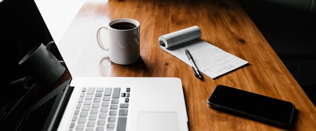 declaração pré-preenchida do IR: imagem de um bloco de notas, caneta, celular, caneca e notebook em cima de uma mesa de madeira