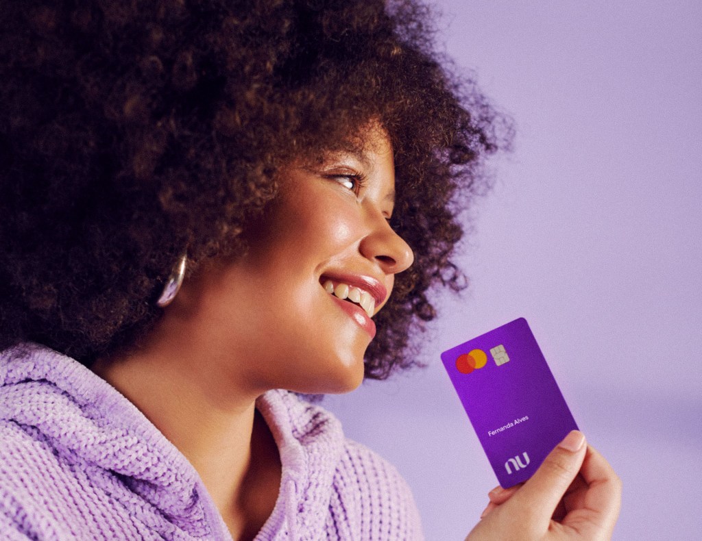 mulher sorrindo, vestindo uma blusa lilás e segurando o cartão Nubank