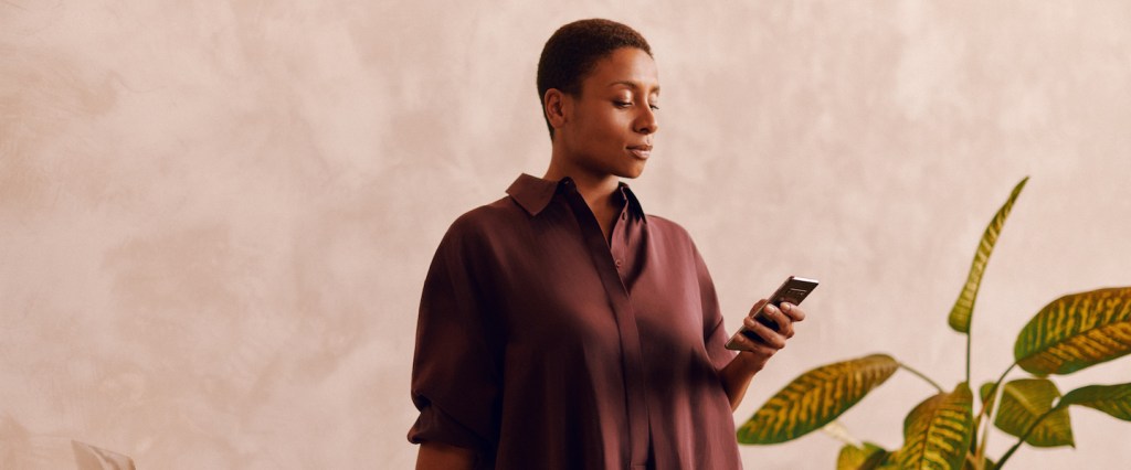 Como receber pagamentos de maquininhas e apps na conta do Nubank: fotografia de uma mulher apoiada com a mão direita em uma mesa e segurando o celular com a esquerda. Sobre a mesa, um computador e vários papeis.