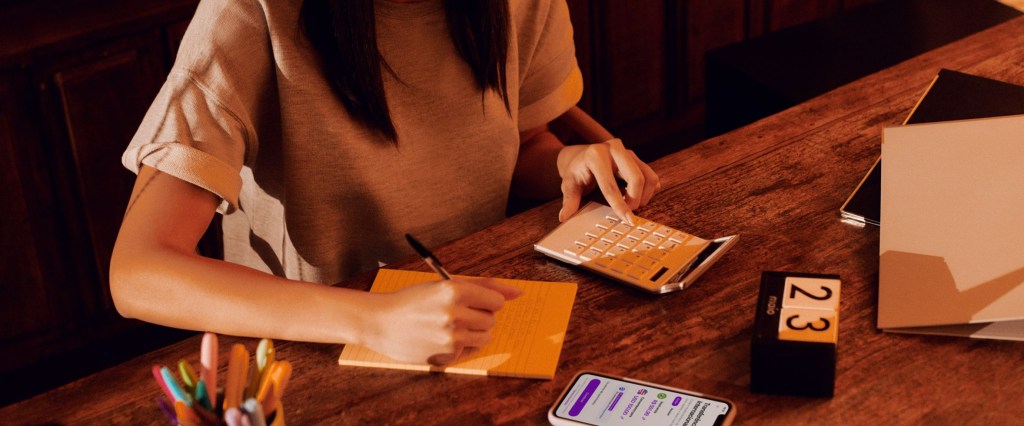 Mulher de pele branca e cabelo comprido está sentada de frente para uma mesa de madeira com um bloco de papel e uma calculadora e o celular onde se lê na tela Transferência Internacional