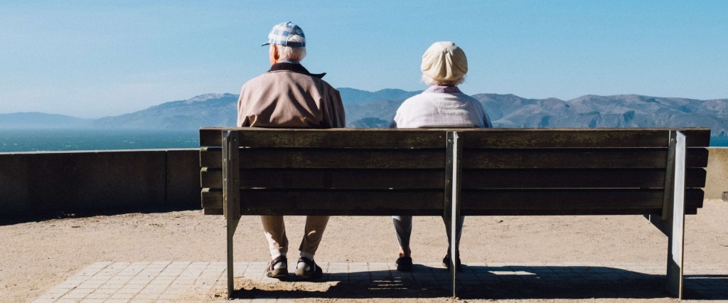 Dois idosos, um homem e uma mulher, fotografados de costas sentados em um banco de madeira, olhando para a frente com um céu azul e montanhas ao fundo