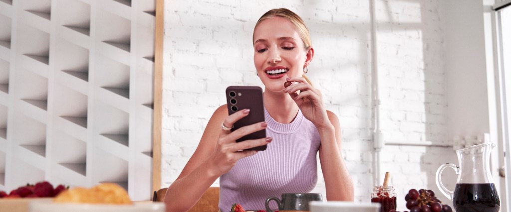 Imagem de uma mulher loira sorrindo, comendo um morango. Ela está de cabelo preso, usando uma blusa regata lilás e segurando um celular em uma das mãos. Ela está à mesa, com várias bebidas e comidas.