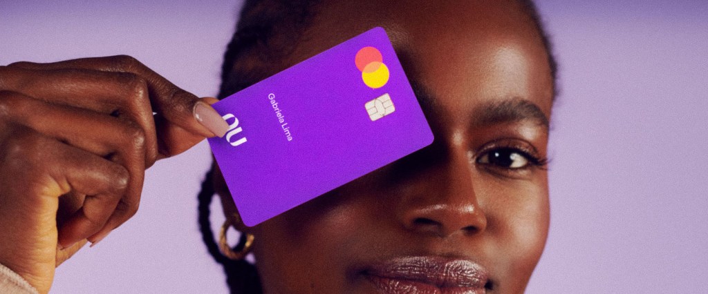 Limite Nubank: retrato de uma mulher olhando para a câmera, com um sorriso pequeno. Ela tem o cabelo curto e com tranças, está vestindo um suéter bege e segurando um cartão Nubank em frente a um dos olhos