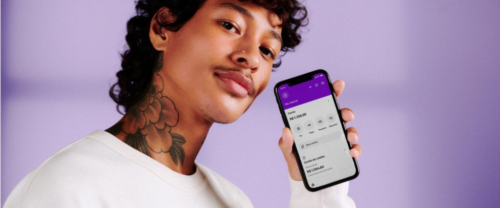 Novo app Nubank 2021: fotografia de homem segurando o celular ao lado do rosto com o novo app do Nubank na tela