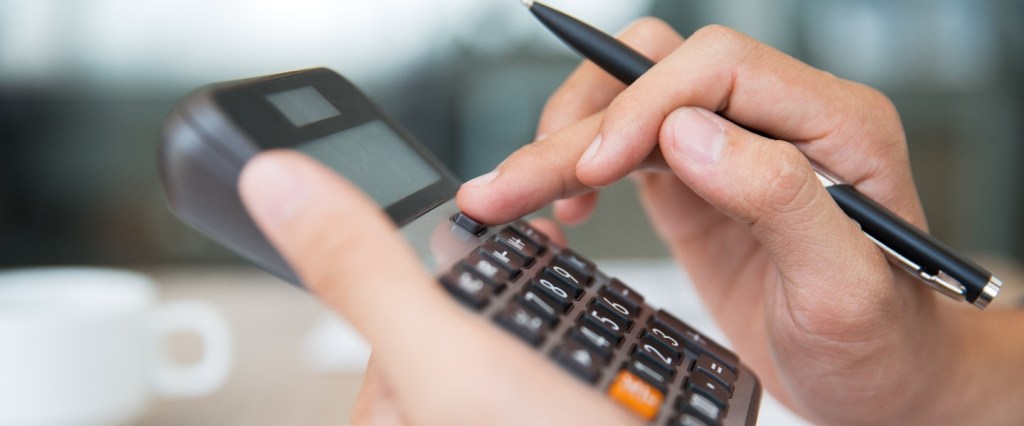 homem faz algum tipo de cálculo segurando uma calculadora com a mão esquerda e uma caneta com a direita. Ao fundo é possível ver uma xícara da café e papeis.