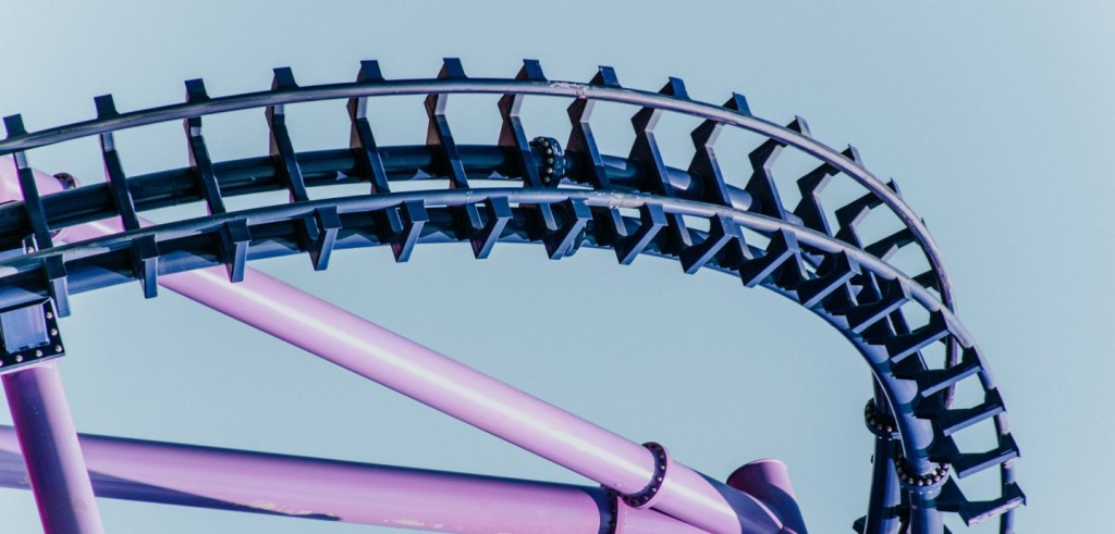 Foto trabalhada digitalmente em tons de roxo, com foco nos detalhes de uma curva da estrutura de uma montanha russa. O objetivo da imagem é ilustrar que mesmo uma estrutura de ferro possui subidas e descidas, assim como a temática abordada no texto, em que as flutuações da renda fixa são atualizadas diariamente pela marcação a mercado.