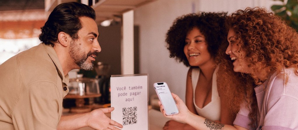 Conta PJ Nubank: imagem de um empreendedor em um café conversando com duas clientes que se preparam para pagar com QR Code