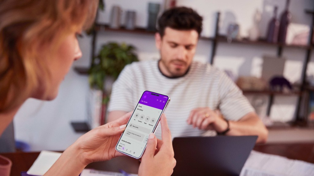 Na imagem aparece, à esquerda, uma mulher branca, ruiva, segurando um celular aberto na tela do app do Nubank. Ao fundo e mais à direita aparece um rapaz branco com uma camiseta branca com listras pretas. Eles estão em uma mesa e ao fundo se vê prateleiras.