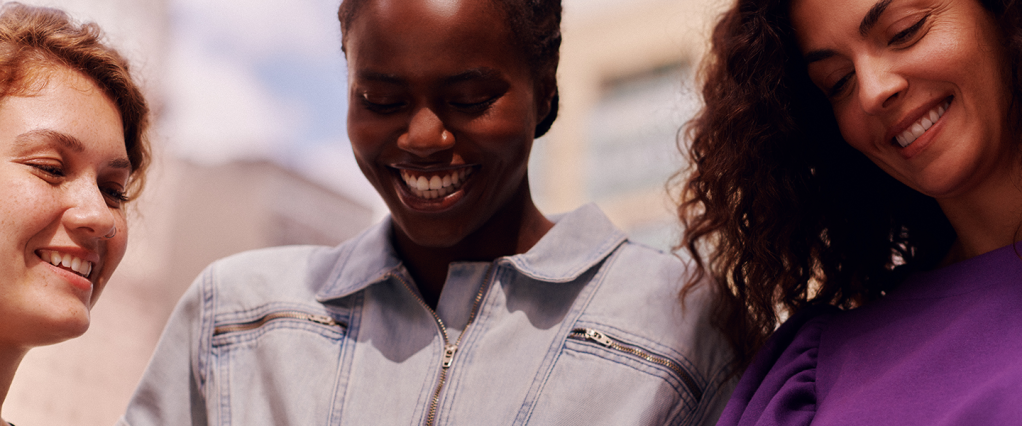 Três jovens sorriem, em uma foto que mostra elas de perto com os rostos ao sol