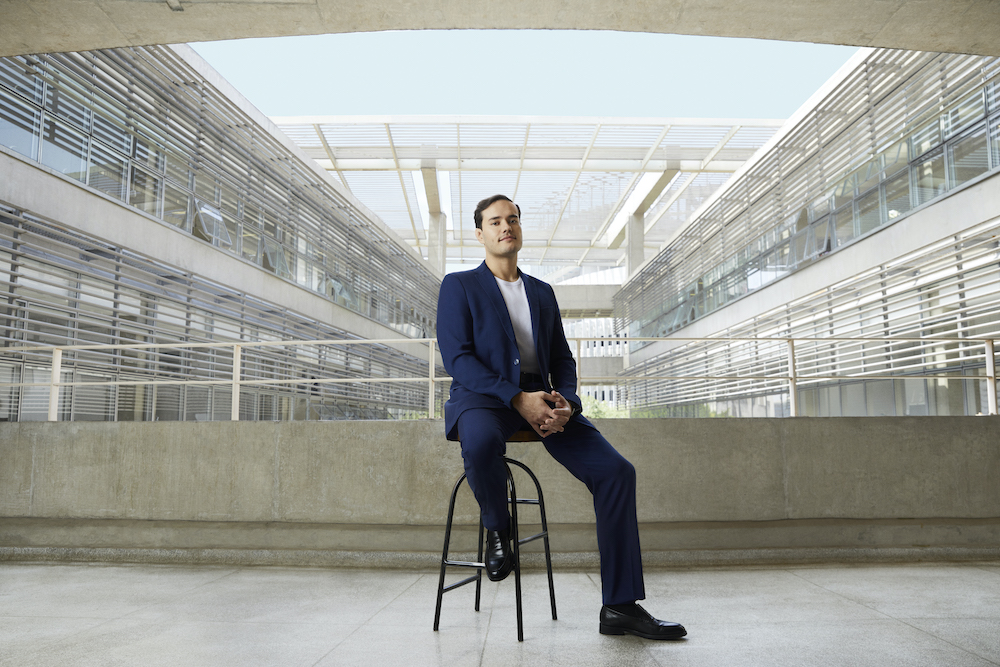 Um homem de terno azul escuro senta em um banco preto. Ele está em um terraço de concreto, com um prédio ao fundo.