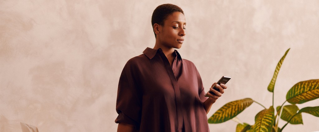 BDR Nubank - mulher de cabelos curtos vestindo uma camisa vinho usa o celular enquanto está em pé ao lado de uma mesa de trabalho; ao fundo existe um vaso de plantas.