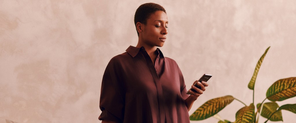 Como receber dinheiro do exterior pelo Nubank? Mulher negra de cabelos curtos vestindo uma camisa vinho usa o celular enquanto está em pé ao lado de uma mesa de trabalho; ao fundo existe um vaso de plantas.