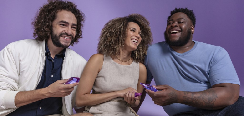 imagem mostra dois homens e uma mulher jovens sorrindo. Eles seguram nas mãos um objeto roxo que representa um pedacinho do Nubank.