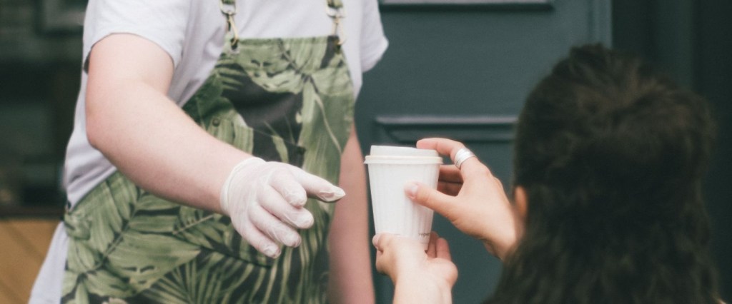 Experiência do cliente: homem de avental serve um café a uma mulher de cabelo es curo curto que está de costas. A foto representa um atendimento. Foto: @DanBurton/Unsplash